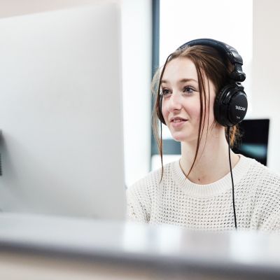 A girl with brown hair and wearing a white jumper sits in front of a big Apple Mac. Her hair is tied up and she is wearing a pair of black headphones.